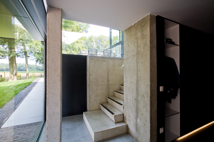 The inside of a house. The staircase is made with concrete and there is a large glazed panel made with glass and aluminium to the side and rear of the room. A lot of natural light is entering the property.