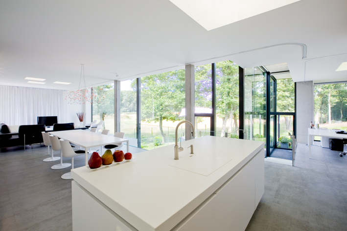 an internal shot of a kitchen. There is a white island in the middle with some fruit on it. To the side of the room, a wall is made with glass and black coated aluminium. You can see trees through the glass. 