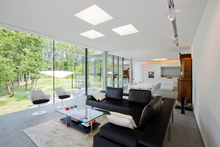 An internal picture of a living space. There is a glazing and aluminium wall to one side which shows a view of the garden. There is a black sofa inside with two white chairs.